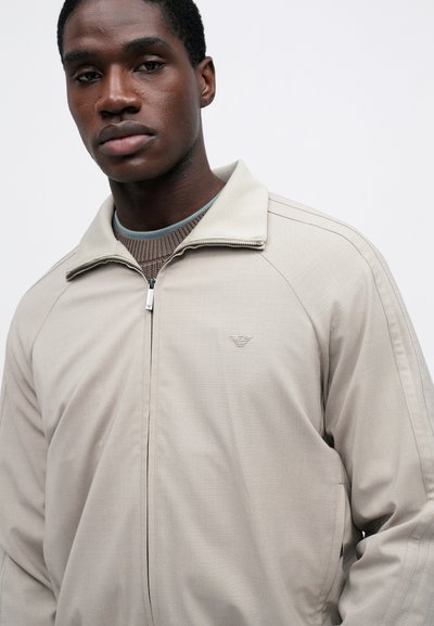 Φωτεινό μπεζ ζακέτα φερμουάρ με ψηλό γιακά, υφή υφάσματος και διακριτή λογότυπο στη θωρακική περιοχή. Διαθέτει ραγλανά μανίκια και λεπτή ραφή.