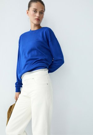 Donna con i capelli raccolti in uno chignon che indossa un maglione blu e pantaloni bianchi, in piedi contro un muro grigio chiaro, con una borsa marrone nella mano sinistra.
