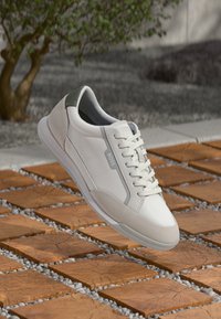 White and beige BOSS sneaker with laces, floating above wooden tiles with small gravel between them outdoors near a tree.