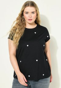 Černé tričko s krátkým rukávem a bílými vzory srdcí rozptýlenými na přední straně. Měkká bavlněná tkanina s kulatým výstřihem.