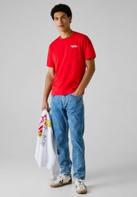 Joven vistiendo una camiseta roja de "Red Bull Racing", jeans azules y zapatillas blancas, sosteniendo una sudadera blanca con logotipos coloridos.