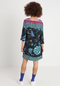 Svart kort klänning med färgglada blommor och fjärils mönster, vida ärmar och en kontrasterande rosa halsringning. Slät tygfinish.