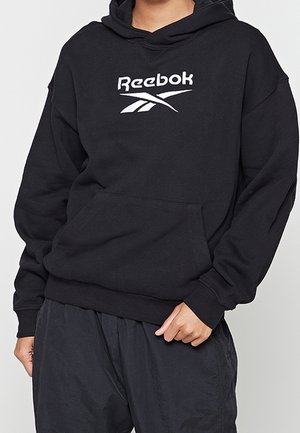 Osoba nosząca czarną bluzę Reebok z białym logo, w zestawieniu z czarnymi spodniami, stojąca z jedną ręką blisko kieszeni.