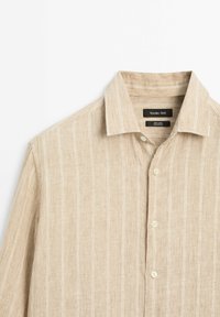 Beige linen shirt with a collar, featuring white vertical stripes and button closure, displayed flat against a neutral background.