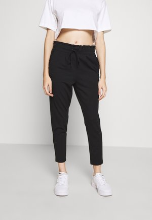 Vrouw draagt zwarte high-waist broek met trekkoord, wit cropped T-shirt en witte platform sneakers, staand tegen een effen achtergrond.