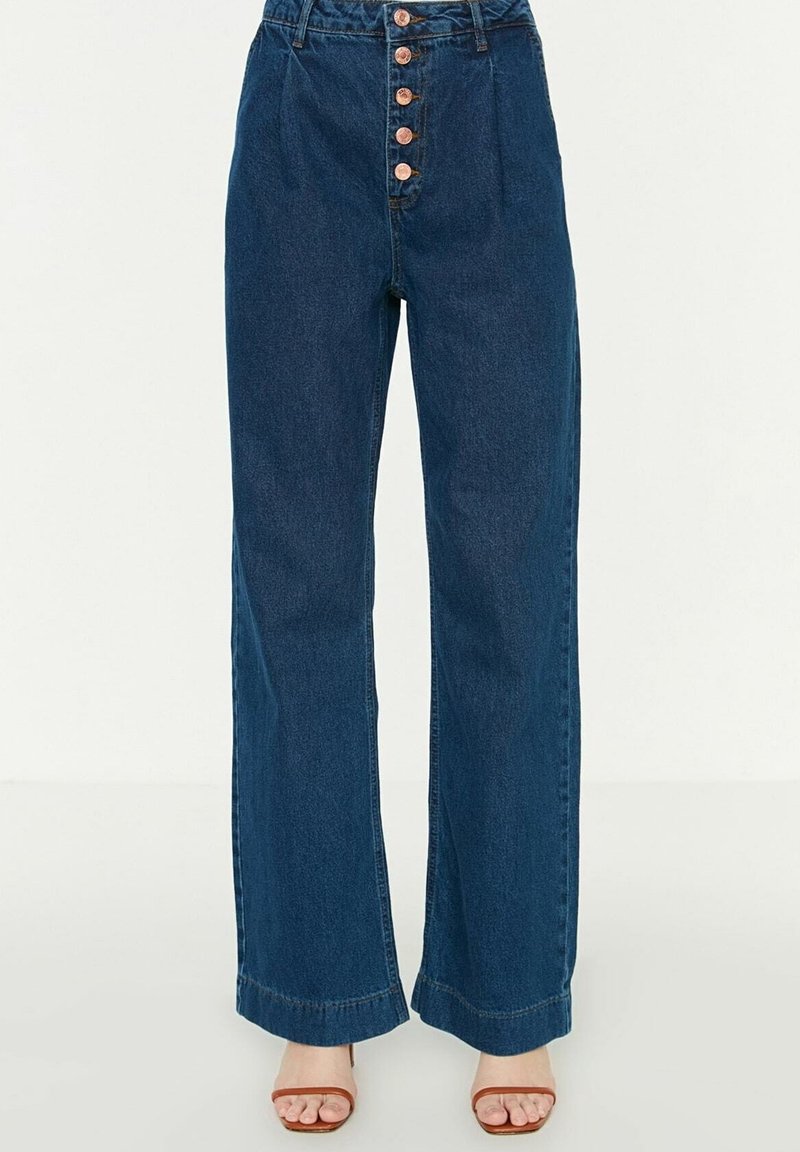 Jeans a vita alta e gambe larghe in denim blu scuro. Presentano sette bottoni di rame lungo la parte anteriore e cuciture visibili all'orlo.