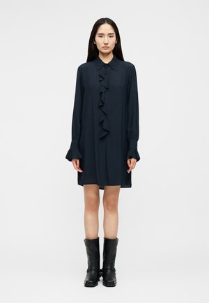 Žena stojaca čelom dopredu, oblečená v čiernych šatách s dlhými rukávmi a volánikmi vpredu, a v čiernych čižmách do polovice lýtok, svetlé jednofarebné pozadie.