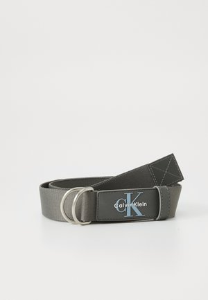 Ceinture en tissu gris avec une boucle double en argent, ornée d'une étiquette noire affichant "Calvin Klein" en blanc et bleu. Surface texturée.