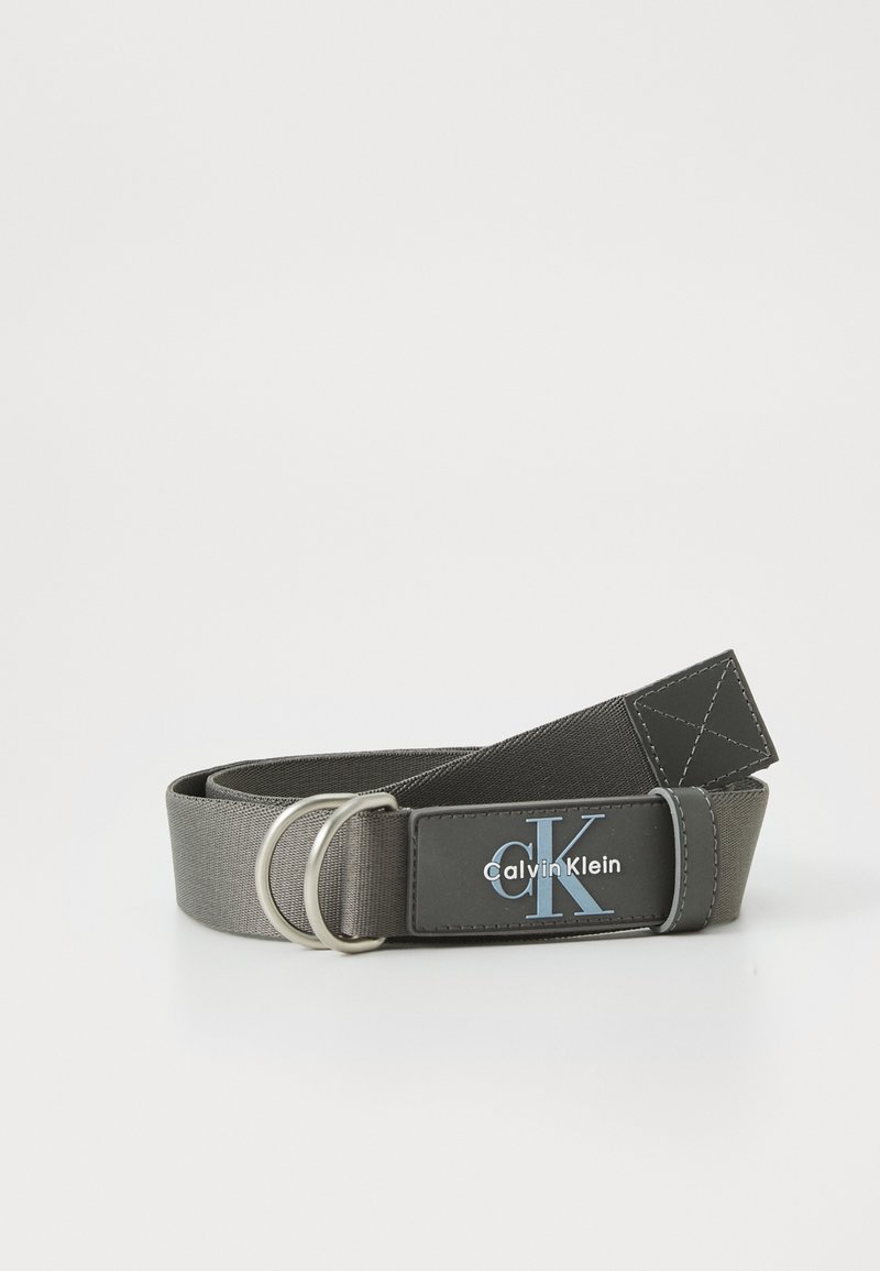 Gray fabric belt featuring a silver double-ring buckle, with a black label displaying "Calvin Klein" in white and blue. Textured surface.