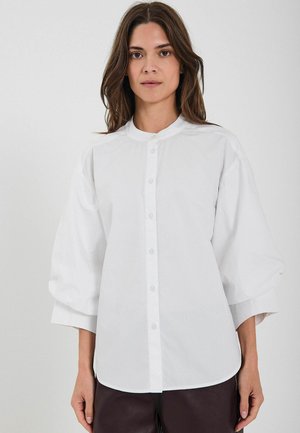 Bela srajca z gumbom in okroglim ovratnikom, z naborkom in širokimi tričetrtinskimi rokavi. Gladko blago in prilegajoč kroj.