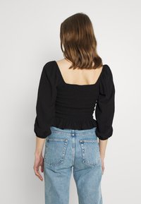 Haut noir smocké avec encolure carrée et manches bouffantes, associé à un jean taille haute bleu clair. Détails visibles de couture et texture du tissu.