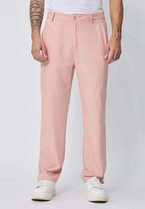 Homme portant un pantalon droit rose clair et des baskets blanches, debout devant un fond clair uni. Tatouage visible sur l'avant-bras gauche.