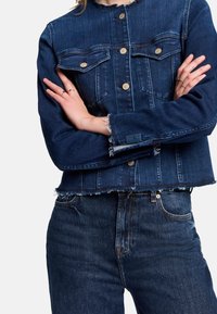 Dunkelblaue Jeansjacke mit tailliertem Design, zwei Fronttaschen und goldenen Knöpfen. Ausgefranste Kanten an den Ärmeln und dem Saum, kombiniert mit passenden Jeans.