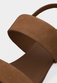 Καφέ σαμουά σανδάλι με φαρδιά λουρί πάνω από τα δάχτυλα και λουρί αστραγάλου σε απλό λευκό φόντο.