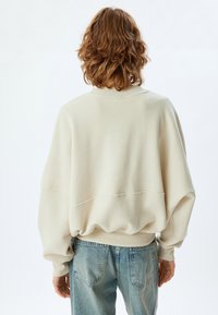 Sudadera de color beige claro, oversize, con un cuello y puños acanalados, que presenta hombros caídos y una textura más suave, combinada con jeans de color azul desgastado.