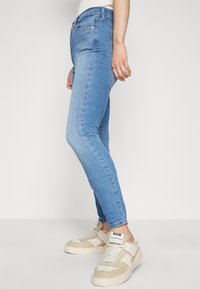 Vaqueros ajustados en denim azul claro, de talle medio, con una textura suave. Combinados con zapatillas beige que tienen un patrón texturizado y un logotipo.