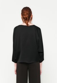 Svart, løstsittende bluse med bred utringning og raglanermer, laget av glatt materiale. Kombinert med brune, vide bukser.