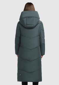 Duga, tamnozelena pufasta jakna s velikom kapuljačom, s chevron šavovima i logom blizu obruba. Mekana, quilted tekstura.