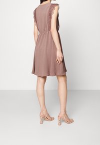 Nainen, joka on pukeutunut mauve-väriseen polvipituiseen mekkoon, jossa on pitsireunukset hihoissa, ja päällään vaaleat, nauhakiilakorkokengät, kuvattuna takaa.