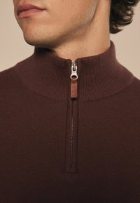Smeđi dukserica na zip, s rebrastom teksturom, visokim ovratnikom i metalnim zatvaračem s kožnom kapicom. Pogodna za svakodnevno nošenje.