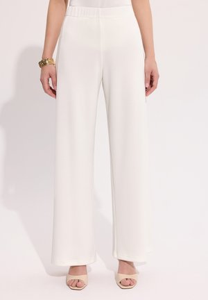 Kobieta nosząca białe, szerokie spodnie z wysokim stanem i elastycznym paskiem, w połączeniu z beżowymi sandałami na odkrytych palcach i złotą bransoletką na lewej ręce.