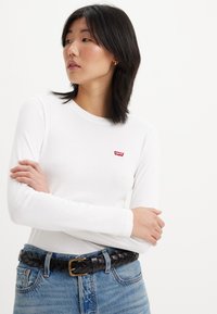 Weißes Langarm-T-Shirt mit Rundhalsausschnitt und kleinem roten Levi's-Logo. Kombiniert mit hellblauen Jeans und einem geflochtenen schwarzen Gürtel.