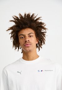 Balta kokvilnas T-kreklā ar pelēku Puma logo un BMW Motorsport zīmolu kreisajā krūtīs. Apaļš kakls, gluda tekstūra.