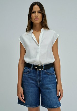 Mujer con cabello largo que lleva una blusa blanca sin mangas metida dentro de unos pantalones cortos vaqueros de tiro alto de color azul con un cinturón negro con hebilla dorada.