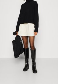 Zwarte cropped trui, crème plissé shorts, zwarte kniehoge laarzen met blokhakken en een getextureerde zwarte tote bag.