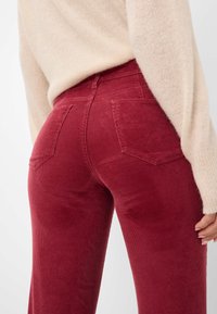 Pantalon en velours côtelé rouge profond avec des côtes verticales, taille haute, poches arrière et une texture douce ; associé à un pull beige clair.
