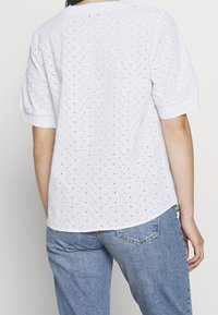 Top bianco ricamato con un motivo testurizzato, maniche corte e orlo arrotondato. Indossato con pantaloni di jeans azzurri chiari.