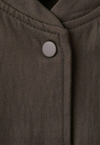 Nahaufnahme eines braunen, gerippten Stoffs mit einem runden, matten Knopf in der Nähe einer Kragennaht.