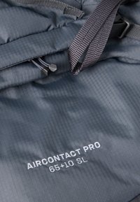 Šedý batoh z ripstop tkaniny s komplikovanými vertikálními liniemi, má nastavitelné popruhy, stahovací šňůrku a text "AIRCONTACT PRO 65+10 SL".