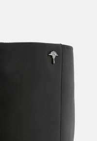 Gros plan sur la tige d'une botte en cuir noir présentant un petit emblème en métal argenté en forme de chardon près du bord supérieur.