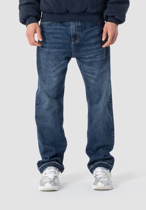 Person trägt dunkelblaue, locker sitzende Jeans, weiße und graue Turnschuhe sowie ein dunkelblaues Sweatshirt und steht vor einem einfarbigen Hintergrund.