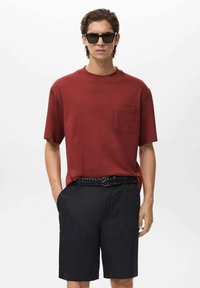 Red short-sleeve t-shirt with a chest pocket, paired with black shorts and a braided belt. Model is wearing dark sunglasses.
