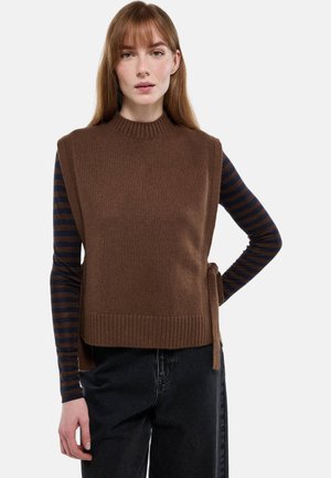 Jeune femme portant un gilet marron en maille sans manches sur un haut à manches longues rayé bleu marine et marron, associé à un jean noir, debout devant un fond uni.