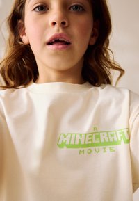 Cremefarbenes Baumwoll-T-Shirt mit einem Aufdruck, der den Text "A MINECRAFT MOVIE" in grünen Blockbuchstaben zeigt und ein Videospiel-Thema hervorhebt.