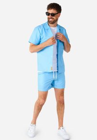 Camicia azzurra a maniche corte e pantaloni corti abbinati, realizzati in un tessuto liscio. Sotto, una t-shirt bianca, abbinata a sneakers bianche e occhiali da sole scuri.
