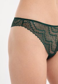 Zielony koronkowy string z falbaniastymi krawędziami i kwiatowym wzorem, eksponujący przezroczystą fakturę oraz niski krój.