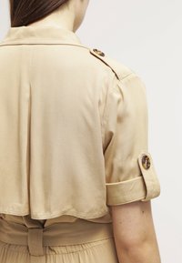 Vue de dos d'une personne portant une veste beige à manches courtes avec des épaulettes boutonnées et une taille ceinturée.