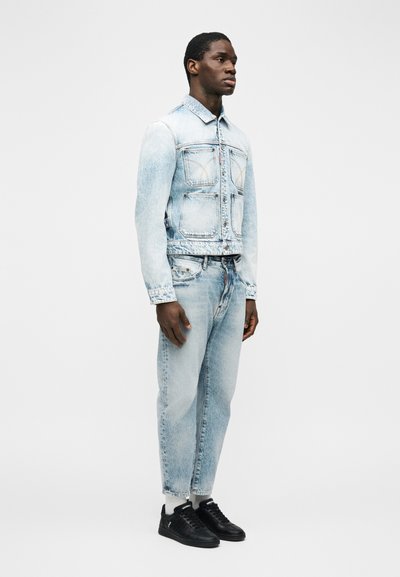 Άνδρας στέκεται πλαγίως φορώντας ανοιχτό μπλε τζιν τζάκετ και τζιν παντελόνι, σετ με λευκές κάλτσες και μαύρα αθλητικά παπούτσια.