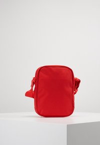 Kompaktní červená crossbody taška s nastavitelným popruhem, obdélníkového tvaru a zipovým uzávěrem, umístěná na bílé podstavě na jednobarevném pozadí.