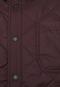 Burgundská prošívaná bunda s černými knoflíky a kapsou na prsou. Má rybí vzor a hladkou, lehkou texturu látky.