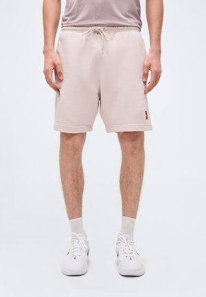 Uomo che indossa pantaloncini rosa chiaro con coulisse, calzini bianchi e sneakers bianche, in piedi davanti a uno sfondo bianco.