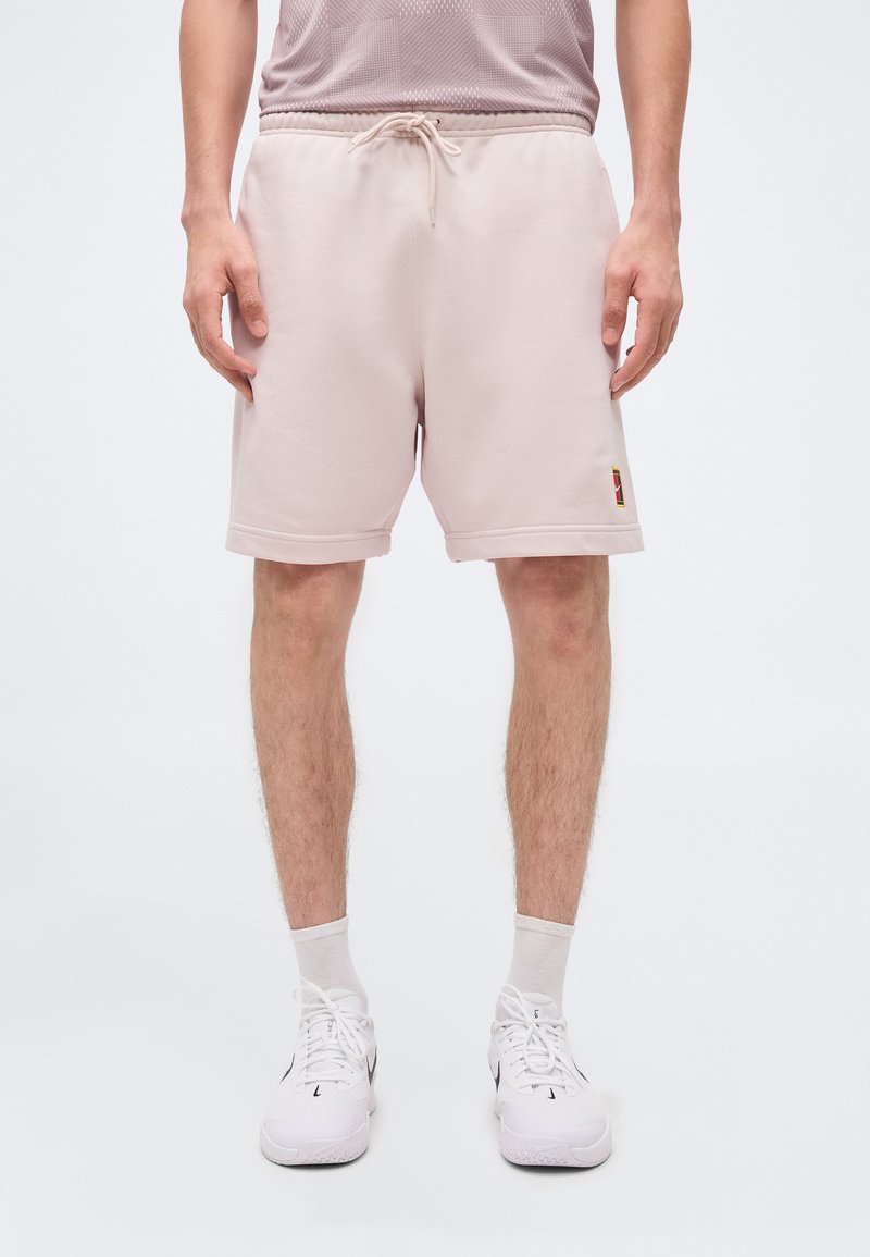 Άνδρας με ανοιχτό ροζ σορτς με κορδόνι, λευκές κάλτσες και λευκά αθλητικά παπούτσια, στέκεται μπροστά σε λευκό φόντο.