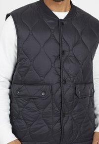 Quilted black vest with a smooth texture, featuring a ribbed neckline, snap buttons, and two front pockets.