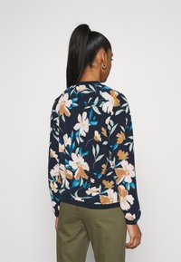 Kvinna med hög hästsvans som bär en marinblå blommig långärmad topp och olivgröna byxor, vänd bort från kameran mot en neutral bakgrund.