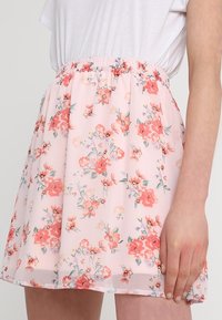 Mini-jupe florale en tissu rose transparent, dotée d'une taille élastique froncée et d'un délicat motif floral rouge et vert sur l'ensemble.