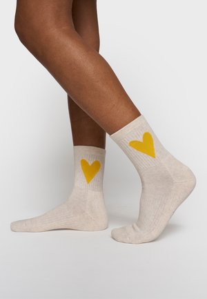 Gambe con calzini beige alla caviglia decorati con cuori gialli, un piede appoggiato a terra, l'altro sollevato sulle punte, su uno sfondo neutro.